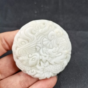 Peut inclure: Un pendentif circulaire en pierre blanche présentant une sculpture en relief complexe représentant un dragon et un phénix. La surface affiche des textures détaillées d'écailles, de plumes et de motifs de nuages tourbillonnants sur le disque.