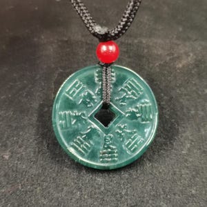 Op de afbeelding: Een cirkelvormige donkergroene hanger met een centrale vierkante uitsparing en in reliëf aangebrachte Chinese karakters. De hanger hangt aan een zwart gevlochten koord met een kleine, gladde, felrode bolvormige kraal boven de hanger.
