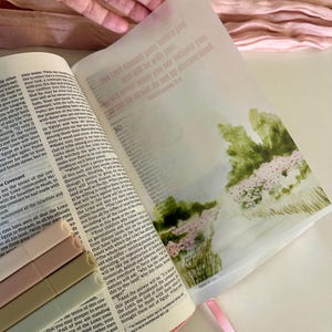 May include: Open Bible with pink ribbon bookmark and a translucent page with watercolor art and text. The text on the translucent page reads "The Lord himself goes before you and will be with you; will never leave you nor forsake you. Do not be afraid; do not be discouraged."