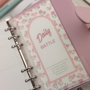 May include: A pink planner with a floral design insert that reads "Daily BATTLE." The insert has a floral border and a Bible verse. The planner is open, revealing the insert and the binder rings.