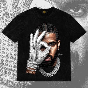May include: A black acid-wash t-shirt featuring a graphic print of a person wearing a sparkling, rhinestone-covered glove over one eye. The design includes a thick, silver-toned chain necklace and the name Drake written in a cursive script.