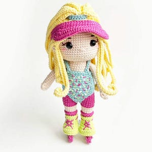 Può includere: Una bambola all'uncinetto con lunghi capelli in filato giallo, che indossa una visiera rosa acceso, un body azzurro con macchioline colorate, pantaloncini rosa e pattini a rotelle verde lime con lacci rosa. La bambola ha occhi neri e pelle chiara.