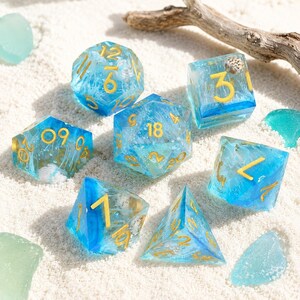 May include: A set of seven polyhedral dice made of translucent blue resin with white swirling inclusions and gold-colored numbers. The dice feature various geometric shapes including a d20, d12, d10, d8, d6, and d4, all displaying clear numerical markings.