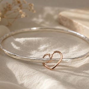 Peut inclure: Un bracelet jonc en argent avec une finition martelée et texturée, orné d'un pendentif en forme de cœur ouvert dans un ton or rose contrastant. Le jonc circulaire est fin et rigide, avec le cœur reposant le long de la circonférence.