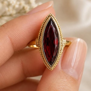 Puede incluir: Un anillo de oro con una gran piedra preciosa de color rojo intenso en talla marquesa, engastada en un borde texturizado similar a una cuerda. La banda de metal es lisa y pulida, sosteniendo la piedra alargada y puntiaguda verticalmente.