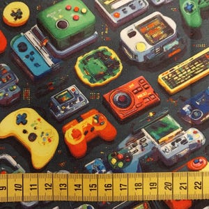 May include: A fabric print featuring a pattern of colourful retro video game consoles and controllers on a dark background, positioned above a yellow measuring tape showing a scale from 9 to 22 cm for size reference of the gaming motifs.