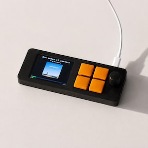 May include: A rectangular black media controller featuring a small square screen displaying the text Aux armes et caetera, four bright orange square buttons arranged in a grid, and a single black circular knob. A white charging cable is plugged in.