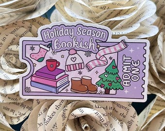 Puede incluir: Una pegatina morada y blanca con el texto "Holiday Season Bookish" y una ilustración de una pila de libros, un gorro de invierno, una bufanda, una taza y un árbol de Navidad. La pegatina también tiene el texto "ADMIT ONE".