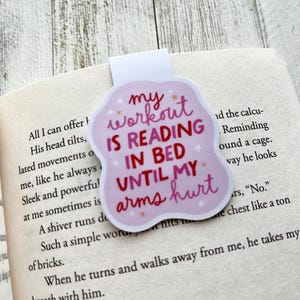 May include: Pink and white bookmark with the text "My workout is reading in bed until my arms hurt".