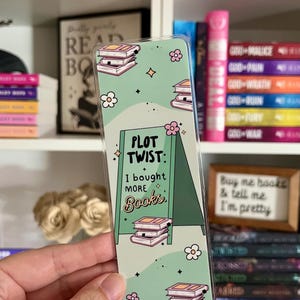 May include: A rectangular bookmark with a light green background featuring stacks of books, small pink flowers, and star icons. The center displays a dark green A-frame sign with the text "PLOT TWIST: I bought MORE Books." in black and brown lettering.