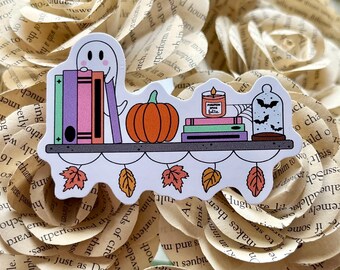 May include: A sticker featuring a shelf with a ghost, books, a pumpkin, a candle, and a cloche with bats. The shelf has autumn leaves hanging below. The candle reads "Pumpkin Spice Latte".