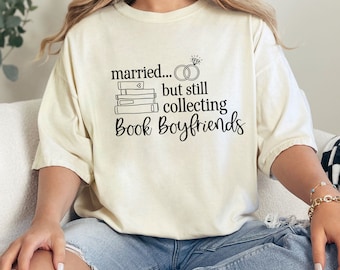 May include: Cream-colored t-shirt with the text "married... but still collecting Book Boyfriends" in black. The design includes a wedding ring, a diamond ring, and a stack of books. The shirt is a casual, oversized fit.