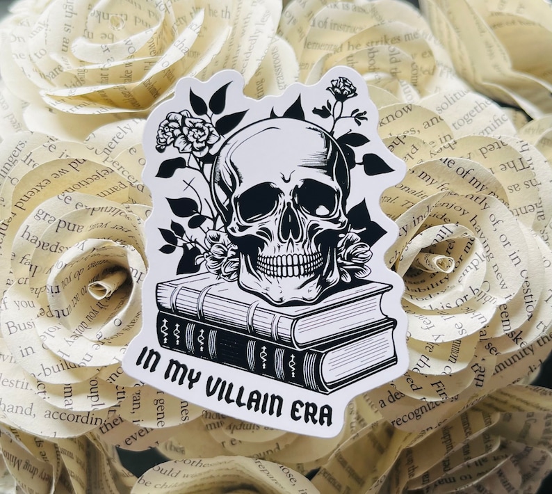 May include: Black and white sticker featuring a skull with a toothy grin, surrounded by flowers and resting on top of two books. The text "In My Villain Era" is printed below the skull.