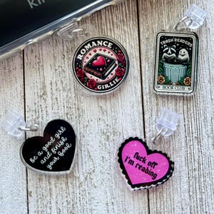 May include: Four different bookish charm designs.  The charms are black and white with white text, pink with white text, and black with white text.  The charms are shaped like hearts and a circle.  The text on the charms reads: "Be a good girl and finish your book", "Romance Girlie", "Trash Readers Book Club", and "Fuck off I'm reading".