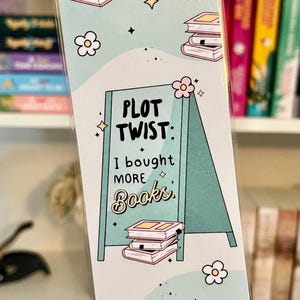 May include: A light blue and green bookmark with a white border. The bookmark features a sign that says "Plot Twist: I bought more books." The sign is in front of a stack of pink books. The bookmark is decorated with pink flowers and stars.