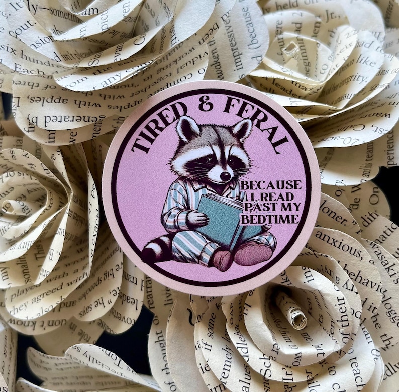 Puede incluir: Una pegatina redonda con fondo rosa y un mapache con pijama leyendo un libro. La pegatina dice "Cansado y salvaje" y "Porque leo m&aacute;s all&aacute; de mi hora de acostarme".