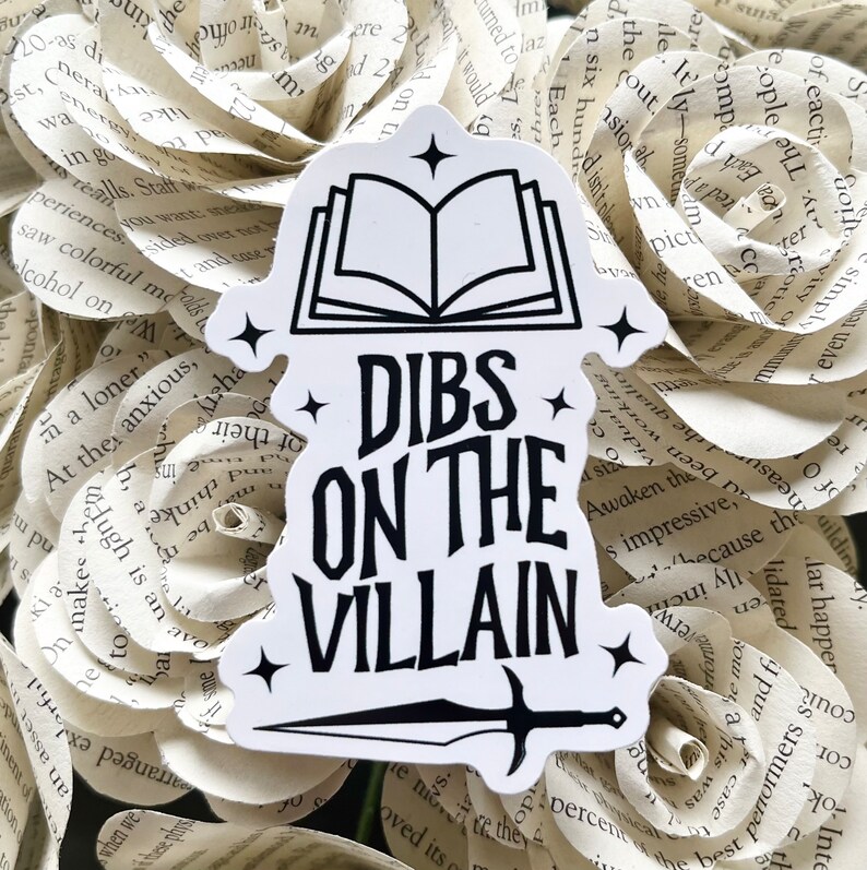 May include: White sticker with black text and a black and white illustration of a book and a sword. The text reads "Dibs on the Villain".
