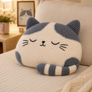 May include: A crochet cat-shaped pillow featuring a cream-colored face and body with dark gray ears, patches, and a striped tail. The cat has embroidered closed eyes, a small nose, and whiskers, creating a soft, plush, and rounded appearance.