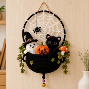 May include: A crochet wall hanging featuring a black cauldron filled with a white ghost in a purple-banded hat, an orange jack-o'-lantern, and a black cat. A white spiderweb is centred above, with a small black spider, green vines, and a mushroom.