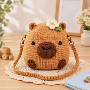 May include: A tan crochet capybara-shaped crossbody bag featuring a dark brown snout, two black bead eyes, and two dark brown feet. A small white flower with a yellow center and green leaves is attached to the top. It has a matching crochet strap.