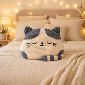 May include: A crochet cat-shaped pillow featuring a cream-colored face and body with dark gray ears, patches, and a striped tail. The pillow has embroidered closed eyes, a small nose, and whiskers, creating a soft, rounded feline appearance.