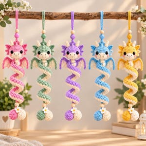 Könnte beinhalten: Fünf hängende Häkel-Drachen-Ornamente in Pink, Grün, Lila, Blau und Gelb. Jeder Drache hat einen geringelten Schwanz, kleine Flügel, schwarze Augen und eine weiße Blumenverzierung an der Basis. Sie sind an passenden Garnschlaufen mit Perlen befestigt.