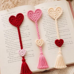 Puede incluir: Tres marcapáginas de ganchillo hechos a mano sobre un libro abierto. Cada marcapáginas presenta un corazón grande en la parte superior, un tallo largo tejido, un pequeño corazón decorativo y una borla en el extremo. Los colores son rojo, rosa claro y crema.