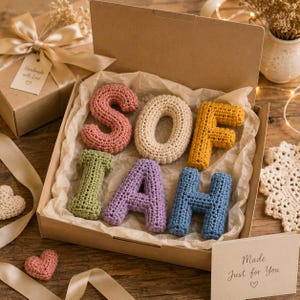 May include: A set of six crocheted letters spelling the name SOFIAH, each in a different color including pink, cream, yellow, green, purple, and blue, arranged inside a cardboard gift box lined with crinkled tissue paper and a gift tag.