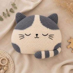 May include: A crochet cat-shaped pillow featuring a two-tone design in dark gray and cream. The pillow has embroidered closed eyes, a small nose, and whiskers. A striped tail in gray and cream is attached to the bottom right side of the pillow.