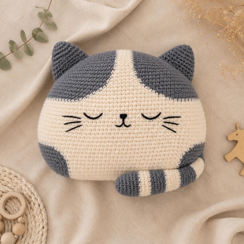 May include: A crochet cat-shaped pillow featuring a two-tone design in dark gray and cream. The pillow has embroidered closed eyes, a small nose, and whiskers. A striped tail in gray and cream is attached to the bottom right side of the pillow.