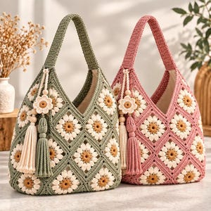 Puede incluir: Dos bolsos hobo de ganchillo, uno en verde salvia y otro en rosa empolvado, con un patrón repetido de cuadrados granny de margaritas con pétalos blancos y centros amarillos. Cada bolso incluye una borla de hilo a juego y un pequeño adorno floral.