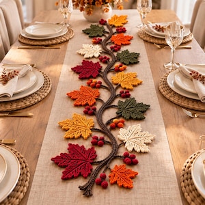 May include: A long, rectangular beige table runner featuring a central, winding brown crocheted vine. The vine is decorated with various crocheted maple leaves in shades of red, orange, yellow, and dark green, interspersed with small red berries.