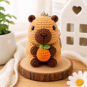May include: A crocheted capybara keychain featuring a light brown body, dark brown paws, ears, and snout, with black bead eyes and pink stitched cheeks. The capybara is holding a small, round, bright orange fruit with two green leaves.