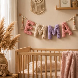 May include: A decorative wall garland featuring individual crocheted letters spelling the name EMMA in shades of pink, cream, and purple. The letters are strung on a white cord accented with wooden beads and small tassels at each end.