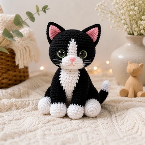 Könnte beinhalten: Ein gehäkeltes schwarz-weißes Katzenspielzeug mit großen grünen Augen, einer rosa Nase und weißen Pfoten. Die Katze hat einen weißen Brust- und Gesichtsfleck, spitze Ohren mit rosa Innendetails und einen Schwanz mit weißer Spitze.