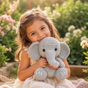 Op de afbeelding: Een grijs gehaakte olifant knuffel met grote oren, een slurf en zwarte plastic ogen. De olifant heeft kleine witte gestikte details op de voeten en een textuur van garen, gemaakt in een zittende houding met afgeronde ledematen.