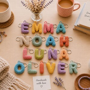 May include: A collection of colorful, crocheted letter keychains arranged to spell the names EMMA, NOAH, LUNA, and OLIVER. Each letter is crafted from yarn in various pastel and bright shades, featuring a metal keyring attached to the top.