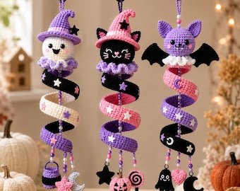 May include: Three hanging crochet ornaments featuring a ghost in a purple hat, a black cat in a pink hat, and a purple bat. Each has a spiral tail decorated with stars, moons, and small charms like a cauldron, pumpkin, lollipop, and ghost.