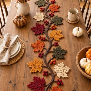 Può includere: Un lungo runner da tavola decorativo caratterizzato da un ramo centrale marrone con foglie d'acero all'uncinetto nei toni del rosso, arancione, giallo, crema e verde scuro, arricchito da piccoli grappoli di bacche rosse lungo tutta la lunghezza.