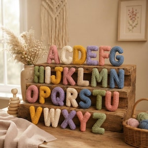 May include: A complete set of twenty-six individual crochet letters from A to Z, crafted in various pastel and vibrant yarn colors. Each letter is three-dimensional and soft-textured, arranged in rows on a tiered wooden display stand for visibility.