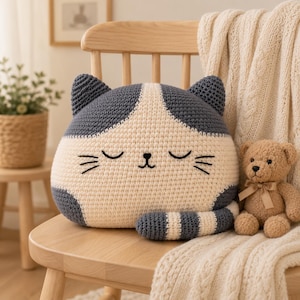 May include: A crochet cat-shaped pillow featuring a cream-colored face and dark gray ears, sides, and tail. The pillow has embroidered closed eyes, a small nose, and whiskers. A small brown teddy bear with a tan ribbon sits next to the pillow.