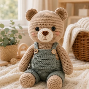May include: A crocheted teddy bear with light brown yarn, featuring black plastic eyes, a dark brown stitched nose, and pink blush accents on the cheeks. The bear wears sage green crocheted overalls with two small wooden buttons on the straps.
