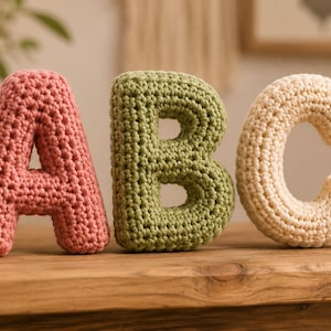 May include: Three plush, stuffed letters A, B, and C crafted from yarn using a crochet technique. The letter A is dusty rose, the letter B is sage green, and the letter C is cream. Each letter has a thick, rounded, and textured surface.