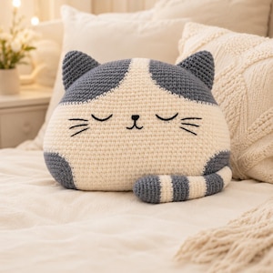 May include: A crochet cat-shaped pillow featuring a cream-colored face and body with dark gray patches on the ears, head, and a striped tail. The cat has embroidered closed eyes, a small nose, and whiskers, creating a soft, plush appearance.