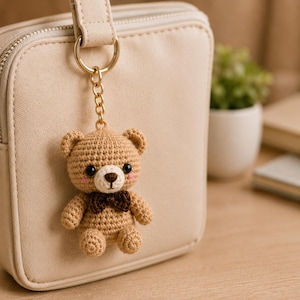 Puede incluir: Un llavero de oso de peluche tejido a ganchillo en color marrón claro con una pajarita marrón oscuro, ojos de plástico negro y nariz bordada. El oso está unido a una anilla y cadena de metal dorado, enganchado a un bolso cuadrado beige.