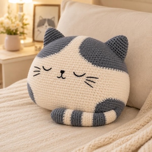 Può includere: Un cuscino rotondo all'uncinetto a forma di gatto con viso e corpo color crema, macchie grigio scuro sulle orecchie e sulla testa e una coda a righe. Il gatto presenta occhi chiusi, un nasino e baffi ricamati con filo nero.
