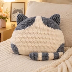 May include: A crochet cat-shaped pillow featuring a cream-colored center and dark gray sides, ears, and a striped tail. The pillow is crafted with a visible yarn texture, showcasing a rounded silhouette with two pointed ears at the top.