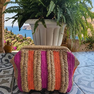 Puede incluir: Un bolso de mano estructurado con un diseño tejido de rayas verticales alternas en rafia color canela natural, hilo magenta vibrante e hilo naranja brillante. El bolso tiene una abertura de marco rígido y una superficie texturizada.
