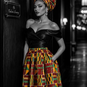 Op de afbeelding: Een zwart off-shoulder topje gecombineerd met een geplooide midi-rok met hoge taille, voorzien van een levendig geometrisch patroon in rood, geel, groen en zwart. Een bijpassende hoofddoek in dezelfde kleurrijke print maakt het geheel af.
