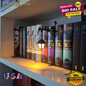 May include: A black metal wall-mounted lantern bookend with a glowing light, positioned between a row of hardcover books on a shelf. The books include titles such as Sherlock Holmes and Binbir Gece Masallari, featuring various spine colors and designs.